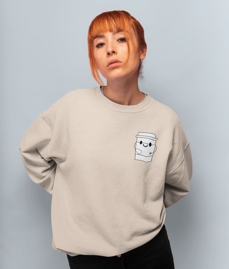 May include: A light beige sweatshirt with a cartoon coffee cup graphic on the left chest. The coffee cup has a smiling face and is holding a small cup.