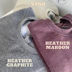 May include: Three folded sweatshirts in different colors: heather graphite, sand, and heather maroon. The sweatshirts are labeled with their respective colors.