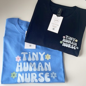 Op de afbeelding: Twee T-shirts met de tekst "Tiny Human Nurse" in een wit, bubbelvormig lettertype met kleurrijke bloemaccenten. Het bovenste shirt is zwart en het onderste shirt is lichtblauw.