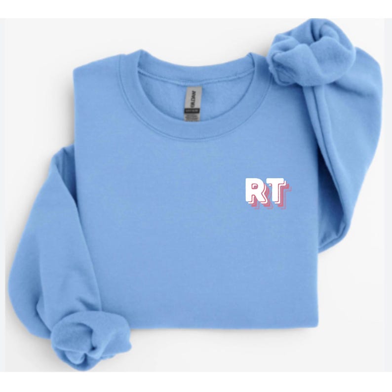 May include: A light blue sweatshirt with the letters "RT" in white with a pink shadow. The sleeves are folded and bunched up on the top right. The sweatshirt is made of a soft material.
