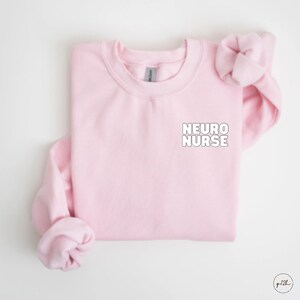 May include: Pink crewneck sweatshirt with the text "NEURO NURSE" in white on the front.