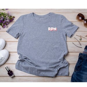 May include: A heather gray t-shirt with the letters "RPN" in white with a red outline. The shirt is displayed on a light wood surface with sunglasses, jewelry, shoes, and a bottle of nail polish.