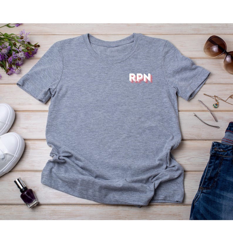 May include: A heather gray t-shirt with the letters "RPN" in white with a red outline. The shirt is displayed on a light wood surface with sunglasses, jewelry, shoes, and a bottle of nail polish.