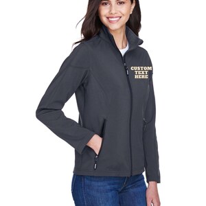May include: A dark grey softshell jacket featuring a full-length front zip, a high collar, and two zipped side pockets. The left chest area displays the text CUSTOM TEXT HERE embroidered in a light yellow font. The jacket is worn over blue jeans.