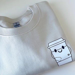 May include: A light beige sweatshirt with a cartoon illustration of a coffee cup with a smiling face. The coffee cup is holding a small cup in its hand.