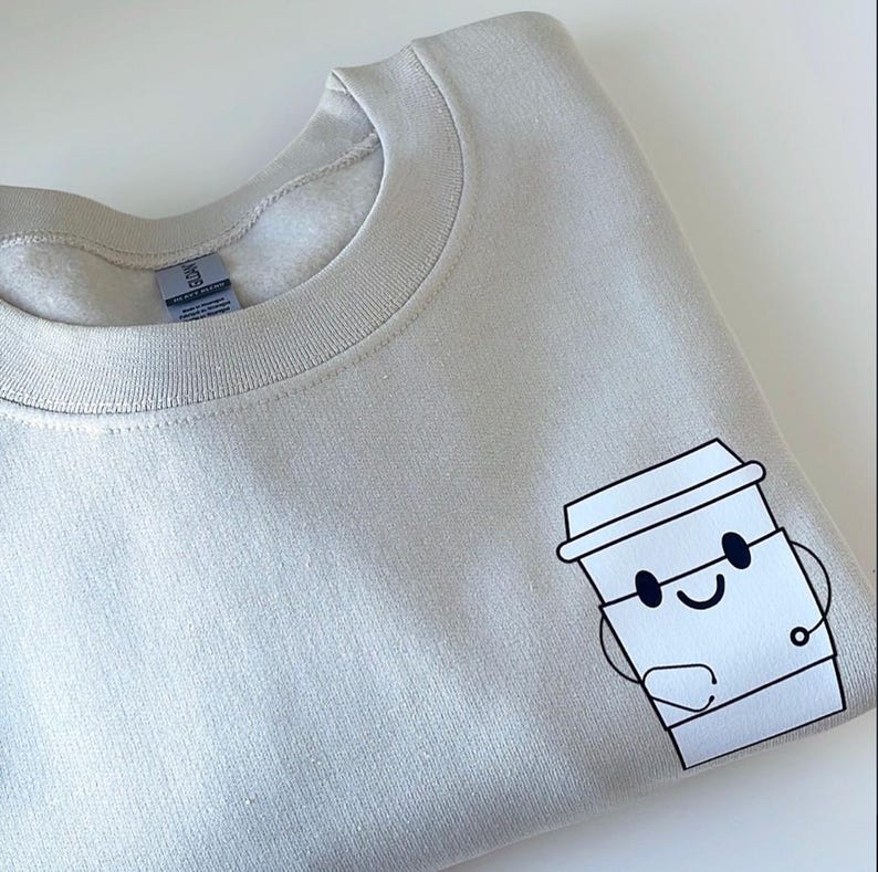 May include: A light beige sweatshirt with a cartoon illustration of a coffee cup with a smiling face. The coffee cup is holding a small cup in its hand.