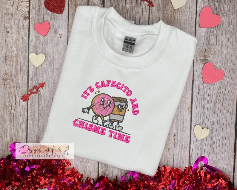 Puede incluir: Sudadera blanca con bordado rosa que dice "It's cafecito and chisme time" con un gr&aacute;fico de donut y taza de caf&eacute;.