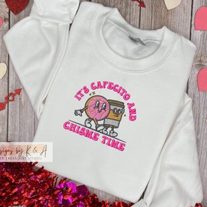 Puede incluir: Sudadera blanca con bordado rosa que dice "It's Cafecito and Chismes Time". El bordado presenta una dona y una taza de caf&eacute; de dibujos animados tomados de la mano.