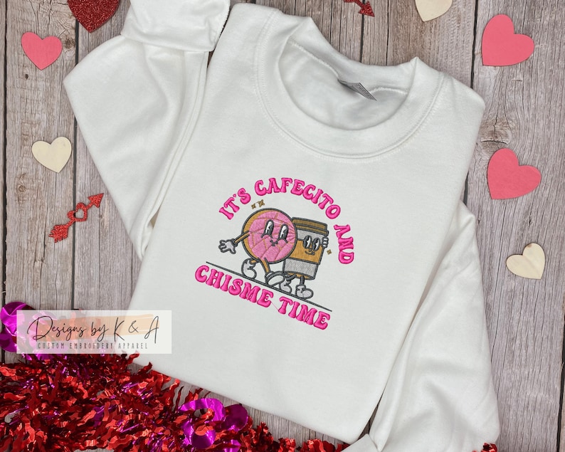 Puede incluir: Sudadera blanca con bordado rosa que dice "It's Cafecito and Chismes Time". El bordado presenta una dona y una taza de caf&eacute; de dibujos animados tomados de la mano.
