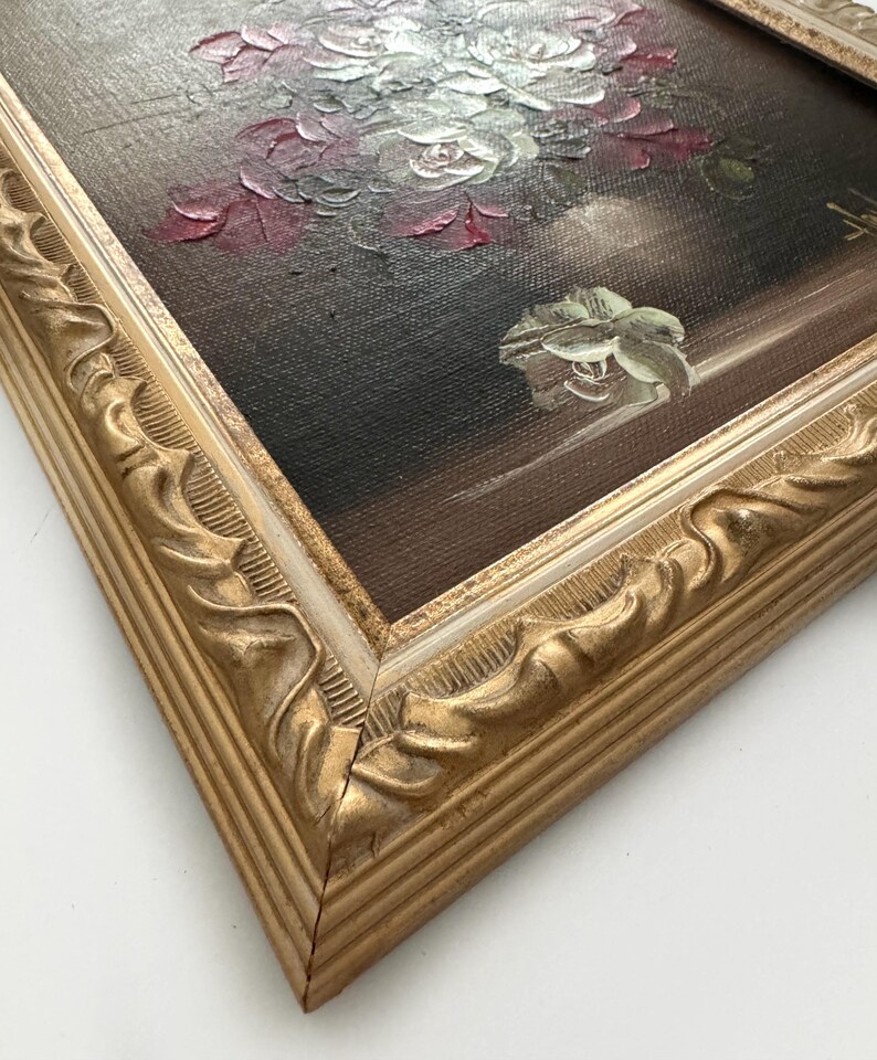 May include: A framed oil painting of a bouquet of white and red roses in a vase. The painting is set against a dark background and has a gold frame with a white inner border.