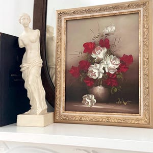 May include: A framed oil painting of a bouquet of red and white roses in a vase. The painting is set against a brown background and has a gold frame.