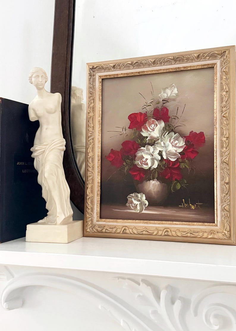 May include: A framed oil painting of a bouquet of red and white roses in a vase. The painting is set against a brown background and has a gold frame.