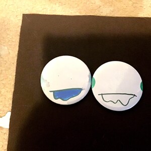 May include: Two white round buttons with different expressions drawn on them. The button on the left has a blue mouth with a drip and the button on the right has a black mouth with a drip.
