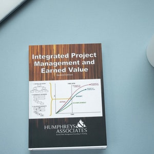 May include: A white book with a brown wooden cover titled "Integrated Project Management and Earned Value" with a graph and a logo for Humphreys & Associates. The book is on a light blue surface with a white coffee mug with black coffee.