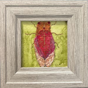 May include: A framed insect art piece. The artwork features a detailed depiction of a red and brown insect with textured wings, set against a textured green background. The frame is a light gray wood.