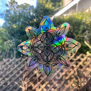 Puede incluir: Una pegatina holográfica con un diseño de mandala en blanco y negro. La pegatina es iridiscente y tiene un efecto arcoíris. La pegatina tiene la palabra "nouvijah" impresa en la parte inferior.