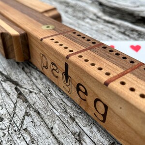 May include: A wooden cribbage board with a brass peg and the word "peg" engraved on the side. The board is made of light and dark wood.