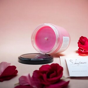 Pode incluir: Uma vela rosa em um pote de vidro com tampa preta. A vela est&aacute; cercada por p&eacute;talas de rosa. O pote tem um r&oacute;tulo branco com texto preto que diz "Soy Wax Candle".