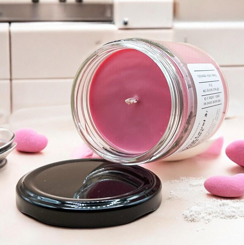 May include: A pink candle in a glass jar with a black lid. The candle is partially melted and the wick is visible. The jar is labelled "Sweetest" and has a small amount of white powder on the side.