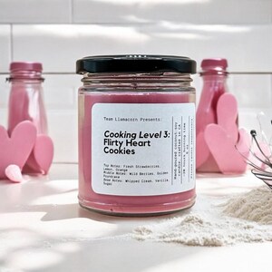 May include: A pink candle jar with a black lid and a white label that reads "Team Llamacorn Presents: Cooking Level 3: Flirty Heart Cookies". The label also lists the candle's scent notes: Top Notes: Fresh Strawberries, Lemon, Orange; Middle Notes: Wild Berries, Golden Poundcake; Base Notes: Whipped Cream, Vanilla, Sugar.