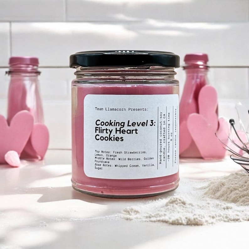 May include: A pink candle jar with a black lid and a white label that reads "Team Llamacorn Presents: Cooking Level 3: Flirty Heart Cookies". The label also lists the candle's scent notes: Top Notes: Fresh Strawberries, Lemon, Orange; Middle Notes: Wild Berries, Golden Poundcake; Base Notes: Whipped Cream, Vanilla, Sugar.