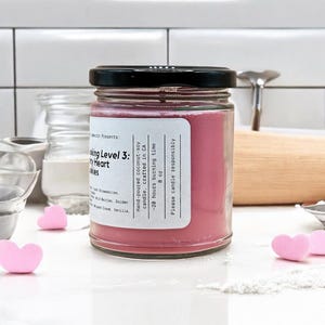 May include: A pink candle in a glass jar with a black lid. The label reads "Baking Level 3: Cherry Heart Cookies" and includes details about the candle, such as the ingredients, origin, and burn time. The candle is 8 ounces and has a burn time of 20 hours.