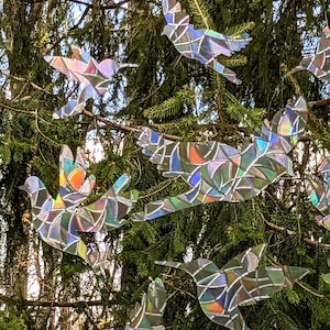 May include: A collection of iridescent, mosaic-like bird cutouts hanging from a tree branch. The birds are made of a reflective material that creates a rainbow effect in the sunlight.
