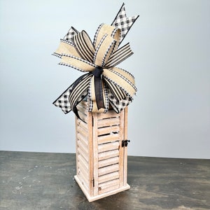 May include: A white wooden lantern with a black and white checkered and striped bow on top. The lantern has a shuttered design and a small door on the front.