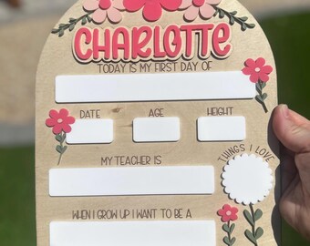 May include: Wooden first day of school sign with the name "Charlotte" in pink and white. The sign has spaces to fill in details like date, age, and height, and is decorated with pink flowers and green leaves.