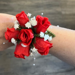 May include: A wrist corsage featuring five vibrant red roses surrounded by small white flowers and green foliage. Delicate white pearl-like beads are interspersed throughout the design, creating an elegant floral accessory.