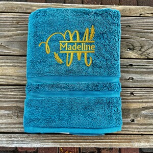 以下が含まれることがあります： 小麦の穂が両側に付いた「Madeline」という名前の金の刺繍のモノグラムが施されたターコイズブルーのタオルです。