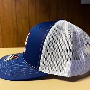 Puede incluir: Una gorra de b&eacute;isbol azul y blanca con un logotipo rojo y blanco. La gorra tiene una parte trasera de malla y una parte delantera s&oacute;lida.