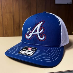 Puede incluir: Una gorra de camionero azul y blanca con una "A" bordada en rojo y blanco y la palabra "The" delante. La gorra tiene una parte trasera de malla blanca y una etiqueta negra y roja que dice "Richardson 1970 Original Trucker 112 Adjustable".