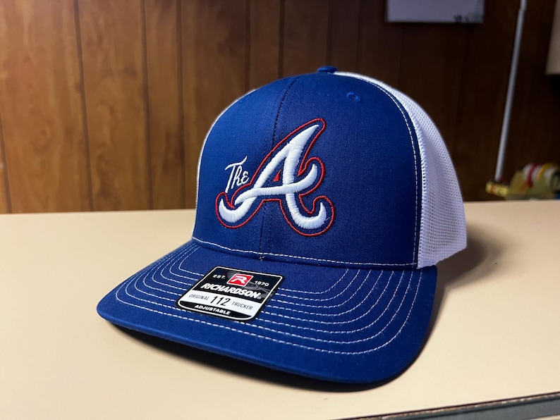 Puede incluir: Una gorra de camionero azul y blanca con una "A" bordada en rojo y blanco y la palabra "The" delante. La gorra tiene una parte trasera de malla blanca y una etiqueta negra y roja que dice "Richardson 1970 Original Trucker 112 Adjustable".