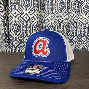 Peut inclure: Une casquette de baseball bleue et blanche avec un logo "A" rouge. La casquette est de style camionneur avec un dos en maille et un devant uni. Le logo est brodé et la casquette est réglable.