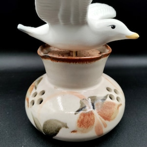 May include: A white ceramic bird figurine perched atop a cream-colored ceramic vase with a brown rim. The vase has a floral design with orange, pink, and green accents and holes for flower stems.