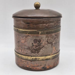 May include: A vintage copper canister with a lid and a small brass knob. The cylindrical container has brass bands around the middle and base. The copper has a weathered, aged appearance, with visible patina and discoloration.