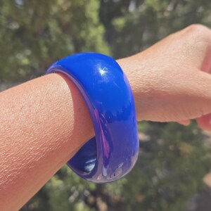 May include: A thick, smooth, and glossy cobalt blue bangle bracelet with a rounded, chunky profile. The surface of the bracelet reflects light, showing a bright white highlight on its curved exterior, creating a solid and bold appearance.