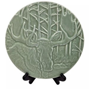 May include: A green ceramic plate with a raised design of a moose in a forest. The plate is on a black stand.