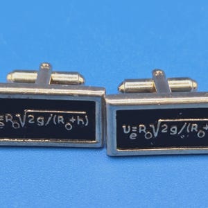 May include: A pair of rectangular cufflinks with a black background and gold-colored trim. Each cufflink features a white mathematical formula. The cufflinks have a gold-colored bar for attachment.