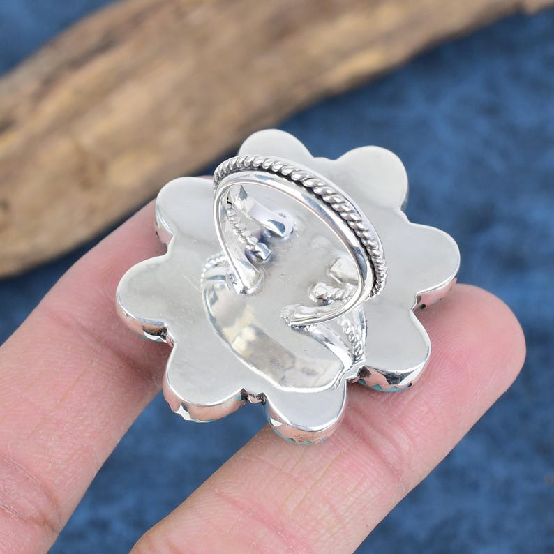 Puede incluir: Un anillo plateado con un engaste grande en forma de flor. El engaste est&aacute; adornado con peque&ntilde;as piedras de color turquesa. La banda del anillo tiene un dise&ntilde;o texturizado. El anillo se muestra desde abajo, revelando los detalles del engaste.