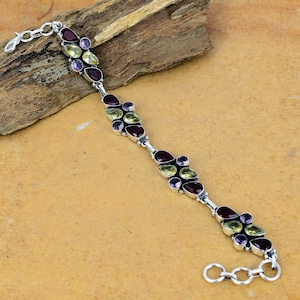 May include: A silver bracelet featuring a series of oval and teardrop-shaped gemstones in shades of red, purple, and green. The gemstones are set in a silver chain, creating a delicate and elegant design. The bracelet is approximately 7 inches long.