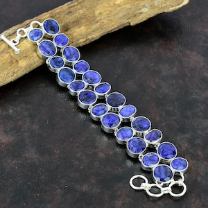 Op de afbeelding: Een zilveren armband met meerdere ovale, diepblauwe edelstenen. De edelstenen zijn gezet in individuele zilveren bezels en verbonden door zilveren schakels. De armband heeft een tuimelsluiting en wordt gepresenteerd op een houten oppervlak.
