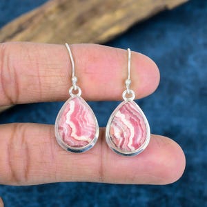 Könnte beinhalten: Ein Paar tropfenförmige Ohrringe mit rosa und weiß gemusterten Steinen, die in Silber gefasst sind. Die Ohrringe hängen an silbernen Haken und zeigen die einzigartigen Farbvariationen innerhalb der Steine. Die Ohrringe werden vor blauem Hintergrund präsentiert.
