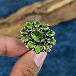 May include: A silver ring featuring a large, oval-cut, olive-green gemstone at the center, surrounded by smaller, round, olive-green gemstones. The ring has an ornate, floral-like design. The ring is held by a hand against a blue and brown background.
