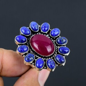 May include: A silver ring with a large, oval-shaped, dark red gemstone surrounded by a circle of smaller, oval-shaped, blue gemstones.