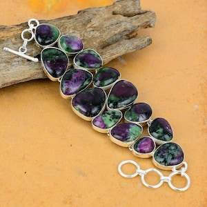 May include: A silver bracelet with a toggle clasp features a row of green and purple gemstone beads. The beads are oval shaped and have a smooth, polished finish.