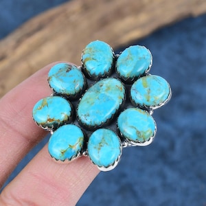 Puede incluir: Un anillo de plata con un grupo de piedras turquesas ovaladas. Las piedras son de un color azul-verdoso vibrante con inclusiones marrones y amarillas. El engaste del anillo tiene un borde festoneado, y las piedras est&aacute;n dispuestas en un patr&oacute;n floral.