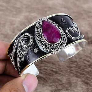 Könnte beinhalten: Ein schwarzes und silbernes Armreif mit einem verzierten Design und einem großen, tropfenförmigen Rubin-Edelstein.