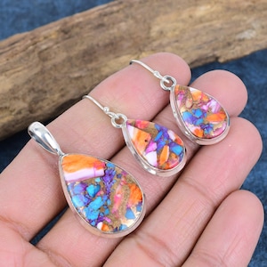 May include: A pendant and earring set with a silver-coloured frame. The teardrop-shaped pieces are filled with a mosaic of orange, pink, blue, purple, and gold-coloured stones. The earrings have a hook closure. The set is displayed on a hand.